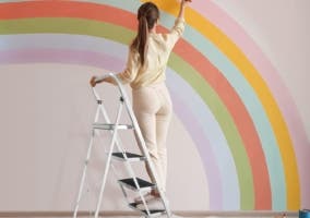 mujer de espalda sobre una escalera multifuncional pintando un arcoiris en la pared